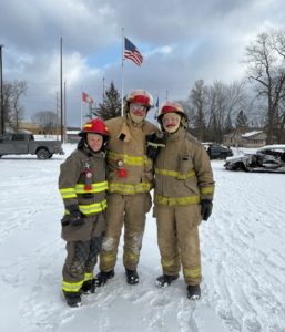 Three people dressed in firefighting gear posing for a photo in the snow with a U.S. flag flying behind them.