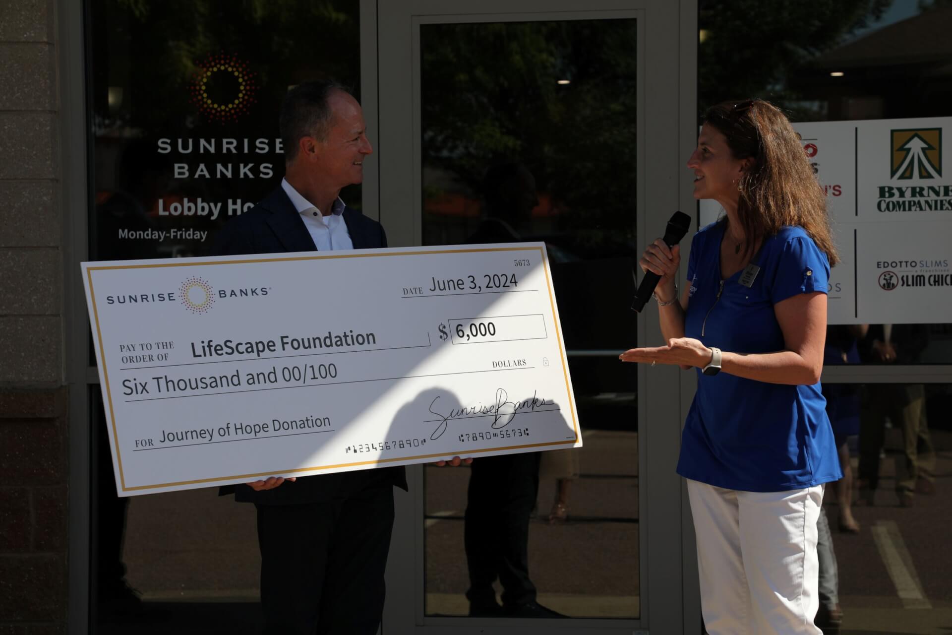 A man holding a giant cardboard check standing next to a woman speaking into a microphone.