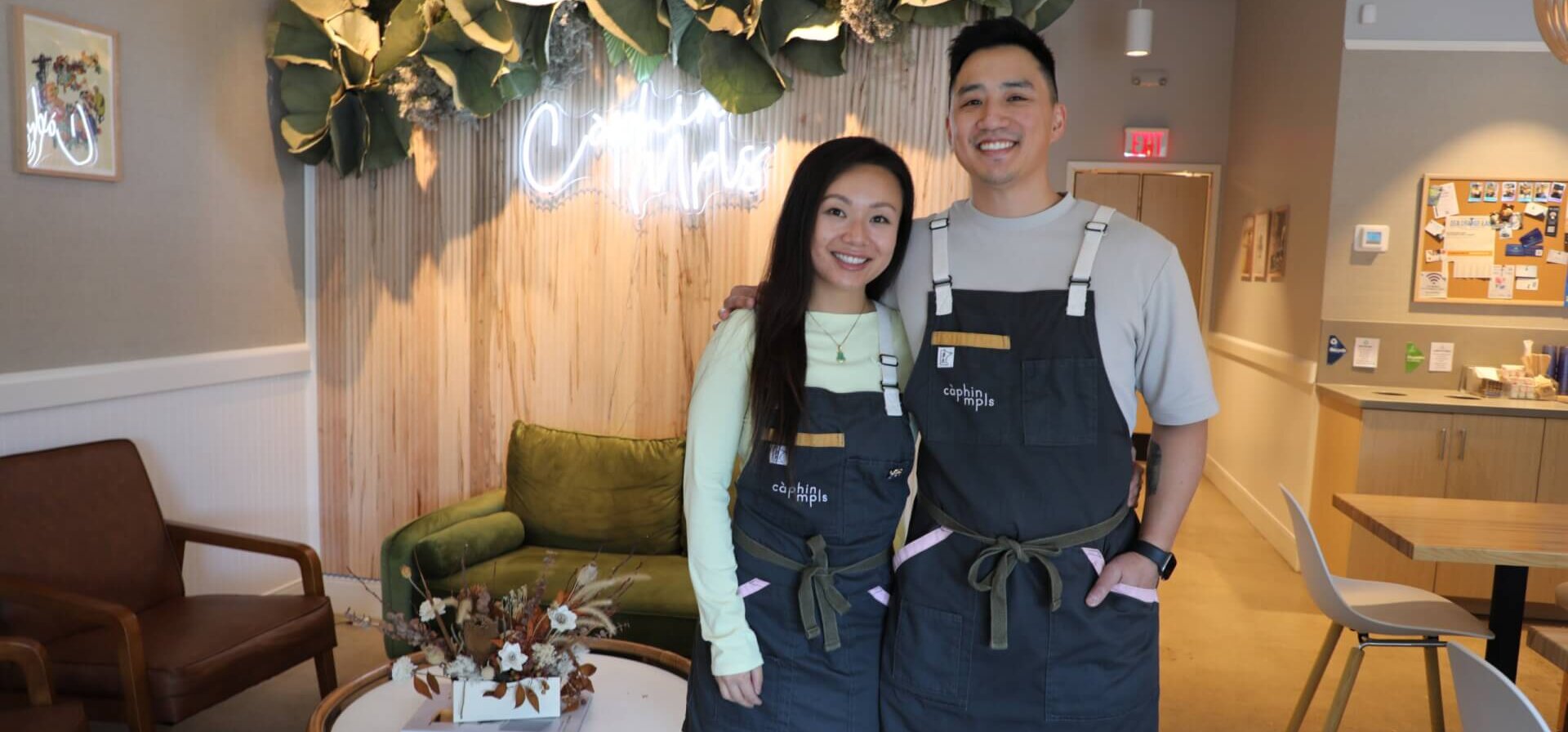 A man and woman standing in front of a neon sign and greenery.