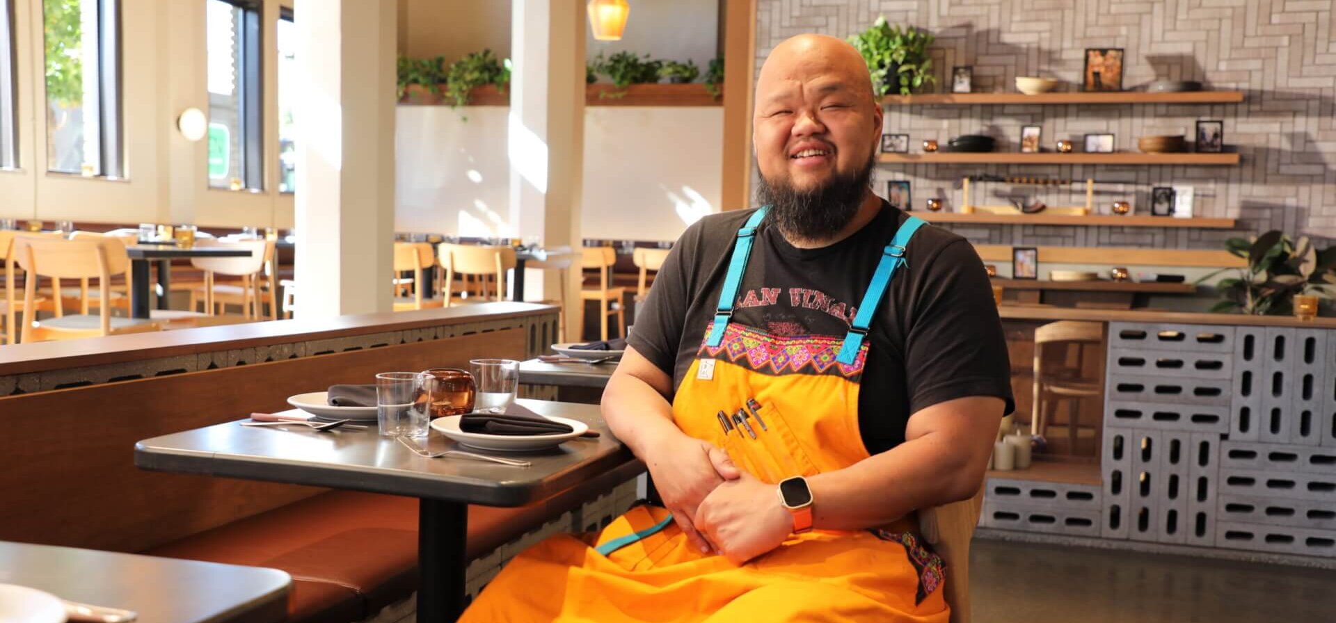 A man sitting at a table in a restaurant wearing an orange chef apron.