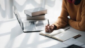 A person writing in a notebook with a laptop and books on a desk.