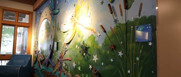 A mural wall depicting a nature scene with grass, water, dragonflies, butterflies and sunlight.