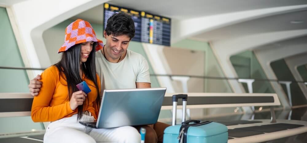 A couple in an airport with luggage sitting down on a bench looking at a laptop set on their laps while one person is holding a credit card
