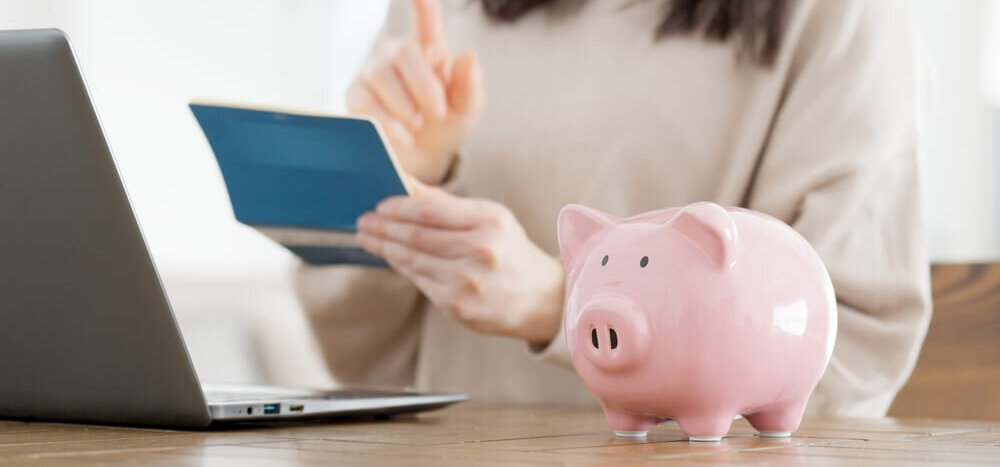 A woman holding a check register with a laptop opened and a piggy bank on a desk