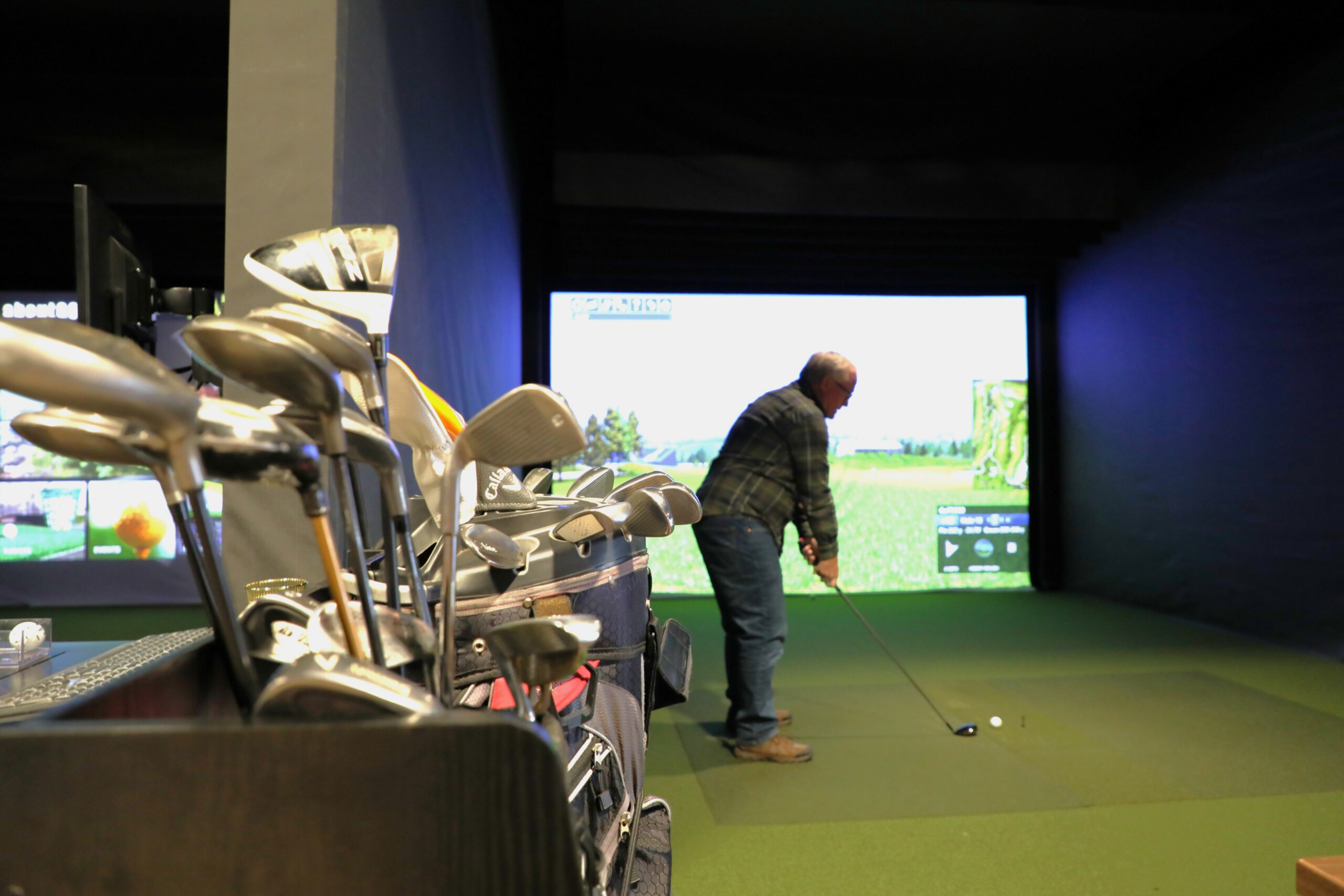 Person swinging a golf club inside an indoor golf simulator, with a set of golf clubs visible nearby.