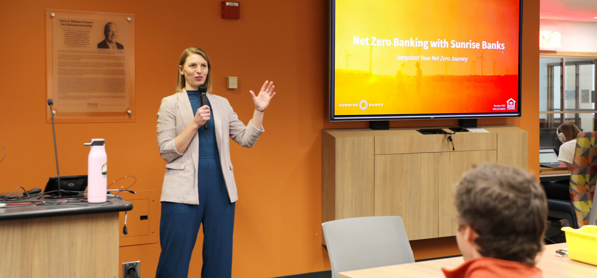 Laura Wildenborg talking to a group of U of M students with a TV screen in the background.