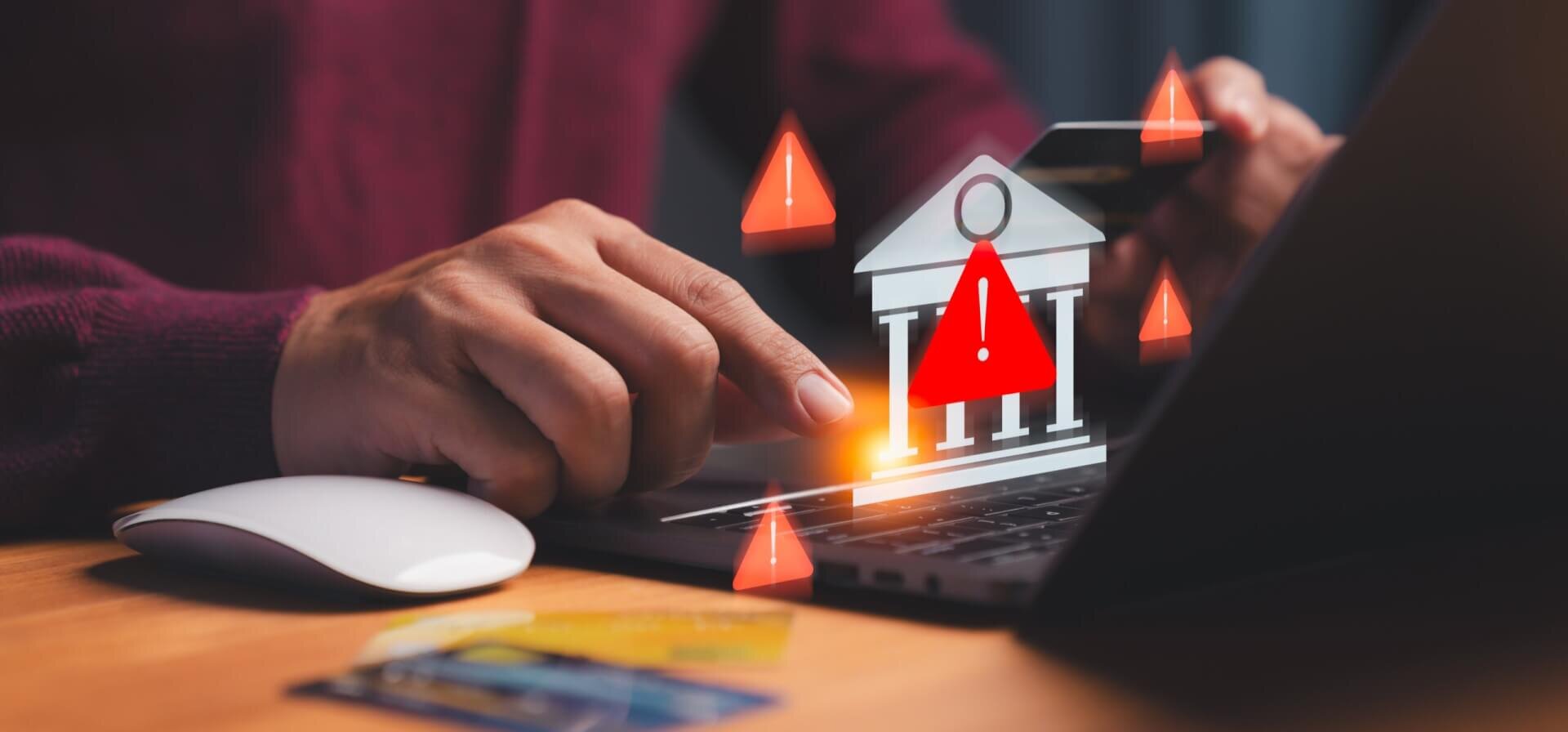 Hands using a keyboard with a bank icon and fraud warning symbols around it.